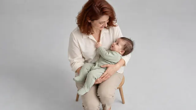 Pencere yakınındaki yumuşak gün ışığında, sade kıyafetleriyle velisinin kucağında kameraya bakan bir bebek